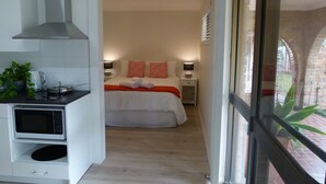 A compact hotel room with a kitchenette, a bed with red and white pillows, and a view of the outside through a window. - The Sanctuary at Paradise Beach (Sanctuary Point)