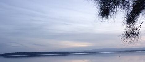 A person performing a balancing act on one leg by a calm lake with a distant shoreline and trees.