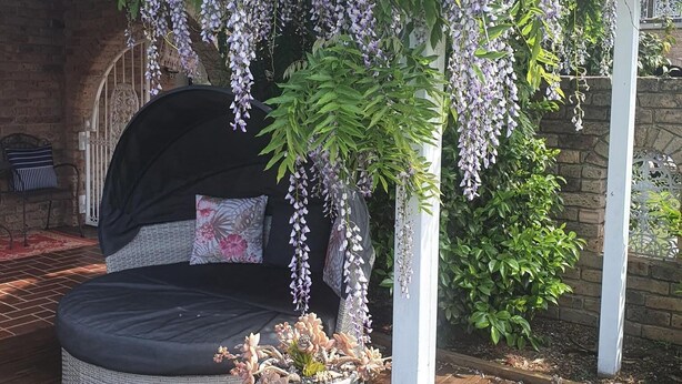 A cozy outdoor seating area with a cushioned bench and a small table, surrounded by a wooden deck and a rock border. - The Sanctuary at Paradise Beach (Sanctuary Point)