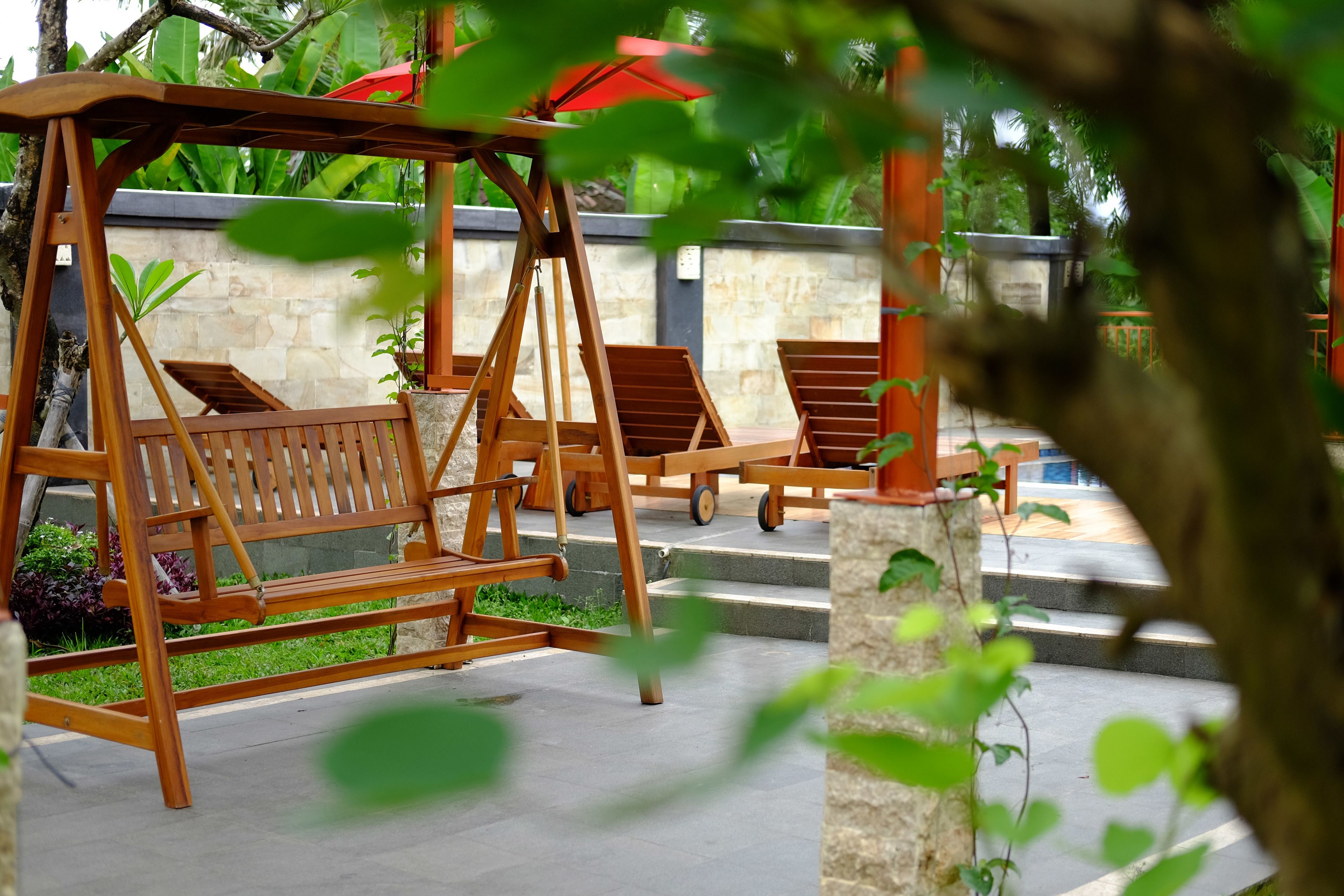 A wooden swing set with two chairs, a stone wall, and a red umbrella in the background.