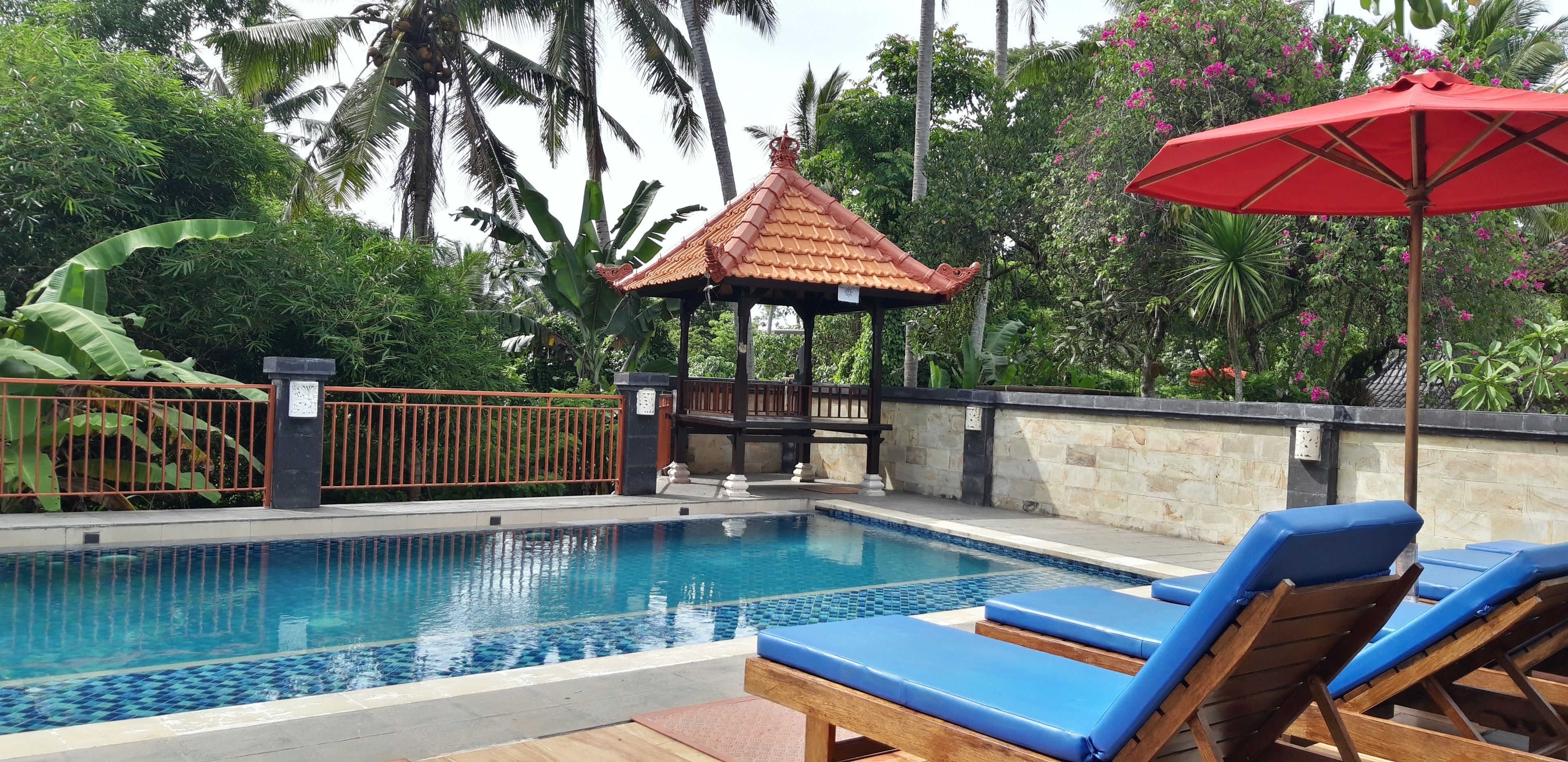 Piscina com bordas em azulejos azuis, rodeada por espreguiçadeiras e um guarda-sol vermelho.