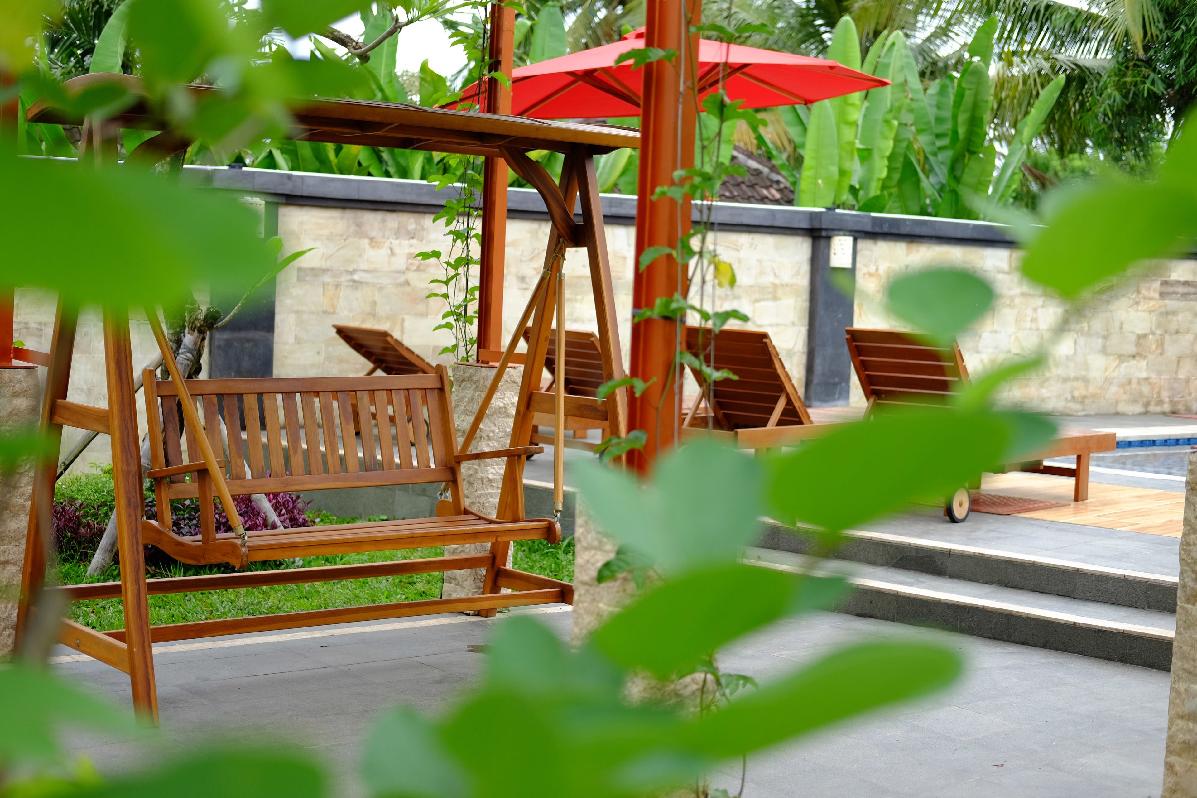 Una silla mecedora de madera, un banco de madera y una zona de piscina con un paraguas rojo.