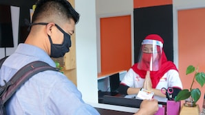 Un mostrador de recepción de hotel con una persona con uniforme rojo detrás, un huésped con camisa azul al frente y un letrero con el nombre del hotel.