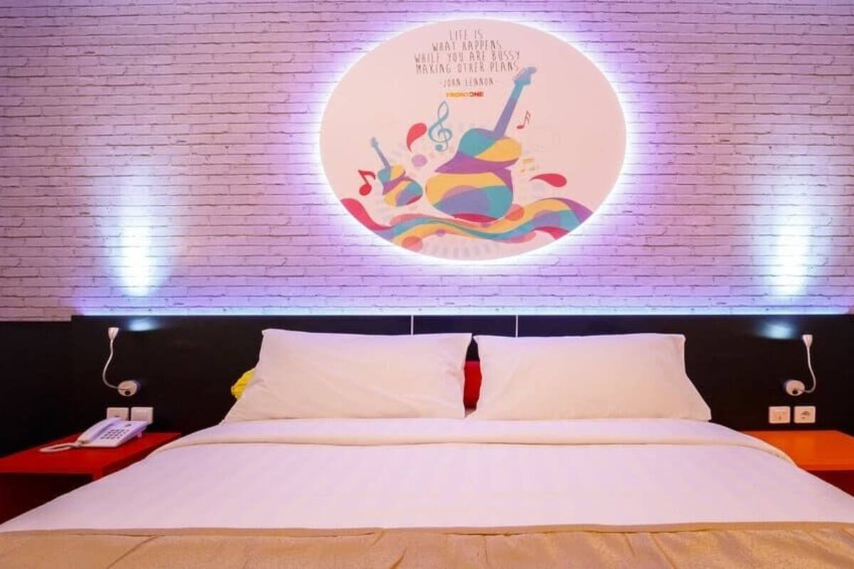 A neatly made bed with white linens, a red nightstand with a phone, and a wall with a circular light fixture displaying a quote.