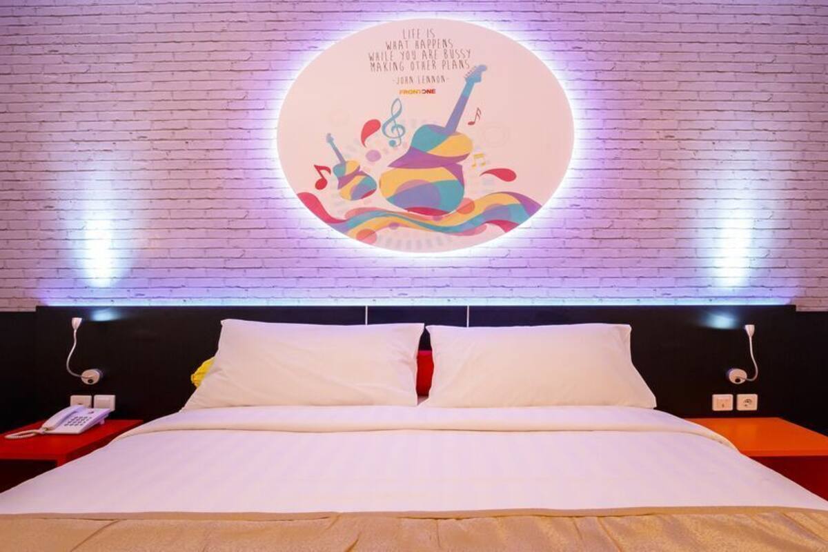 A bed with white bedding, a red bedside table with a phone, and a wall-mounted circular light fixture with a quote.