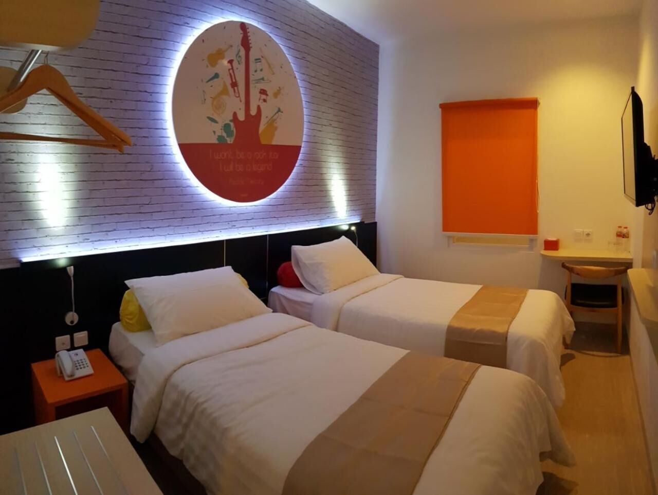 A hotel room with two beds, a wall clock, a bedside table with a lamp, and a small desk with a chair.