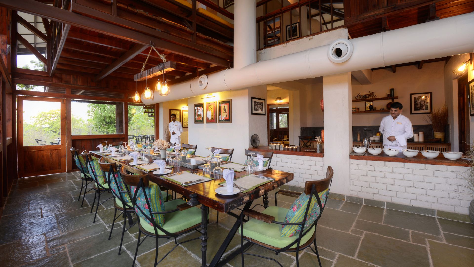 A dining area with a long wooden table set for a meal, surrounded by green cushioned chairs.
