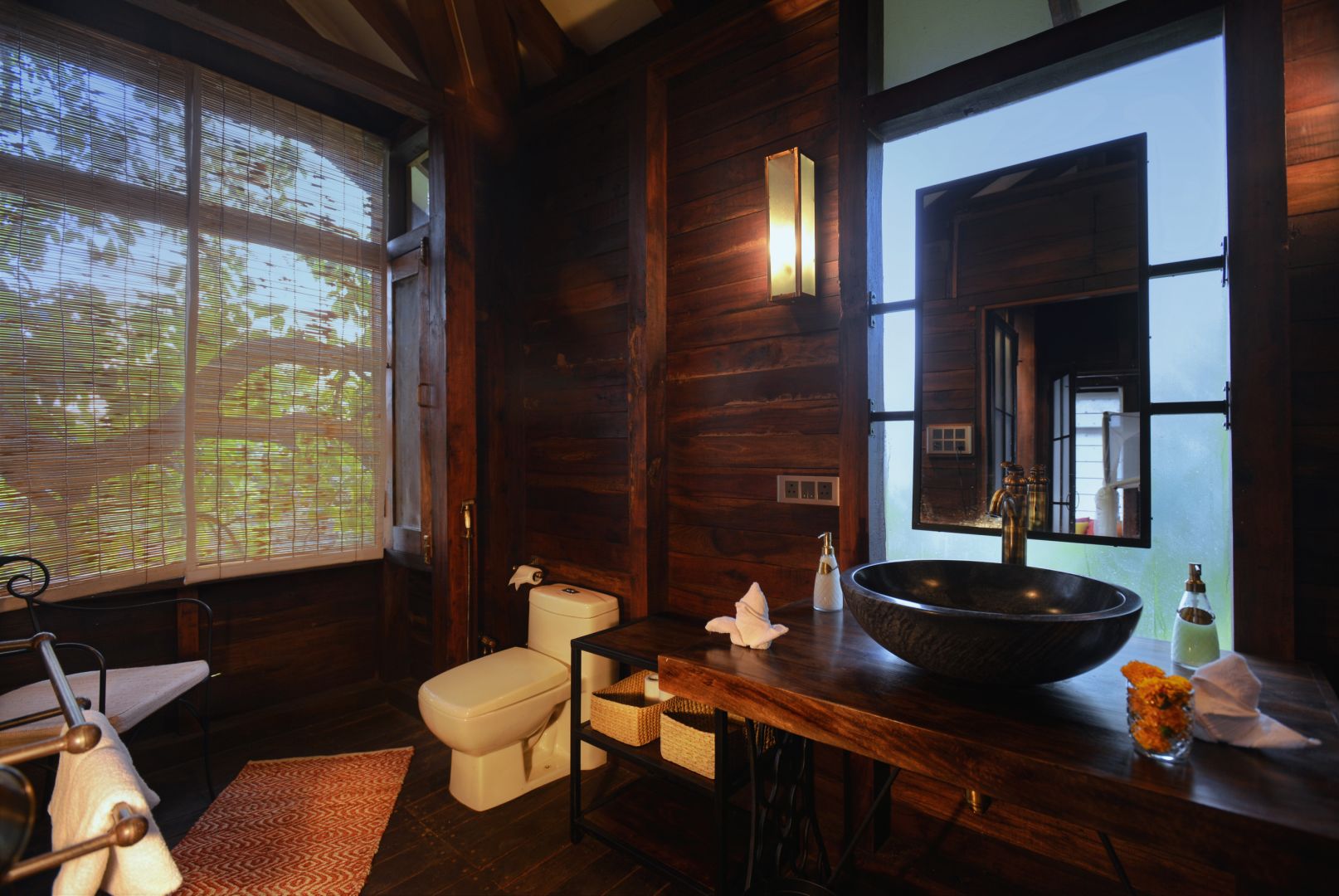 A wooden cabin bathroom with a large window, a white toilet, a wooden vanity with a large bowl sink, and a mirror.