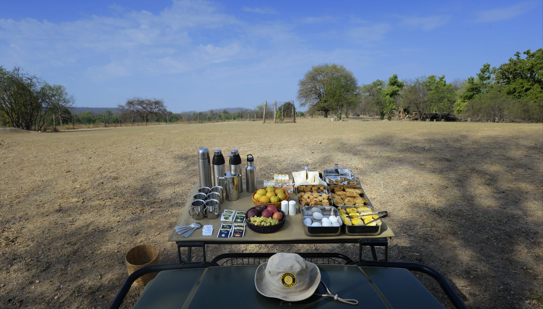 A picnic setup in a dry, open field with a table laden with food, drinks, and utensils.