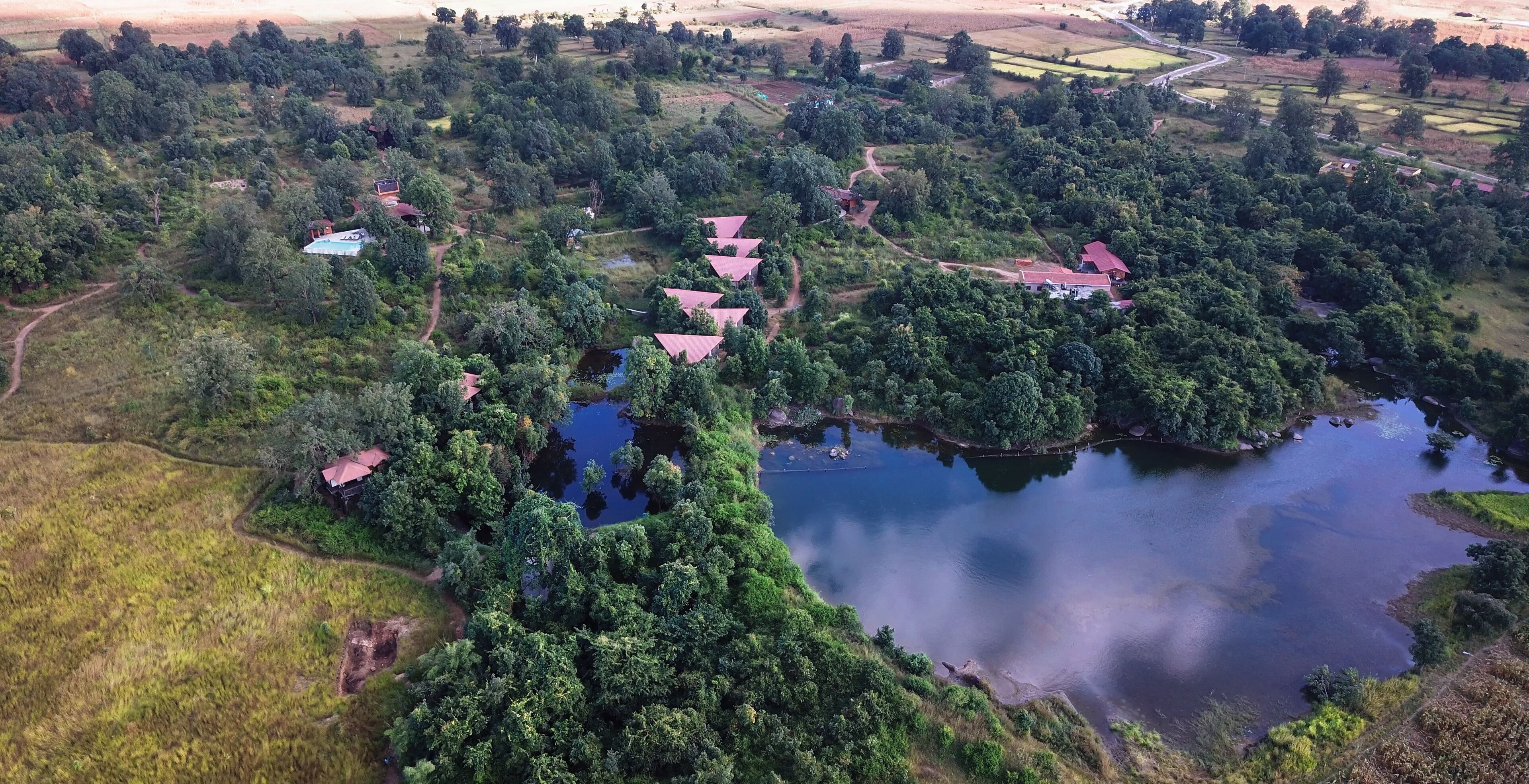An aerial view of a rural area with a lake, surrounded by dense greenery and several buildings with red roofs.