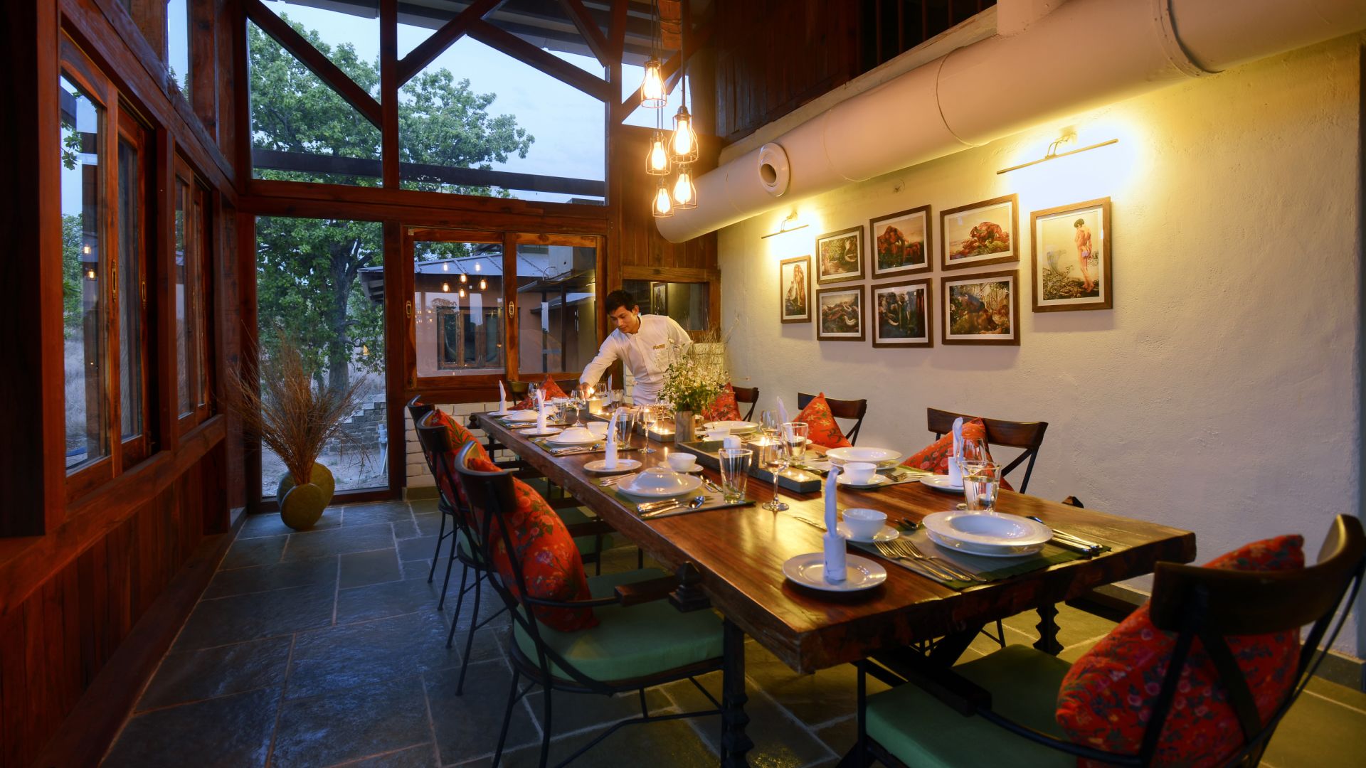 A dining area with a long wooden table set for a meal, surrounded by chairs with red cushions.
