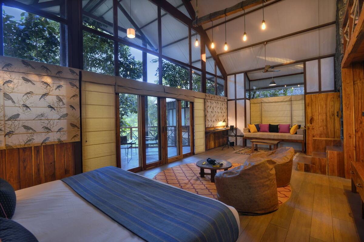 A spacious room with a large bed, a sofa, and a dining area. There are large windows and a view of trees outside.