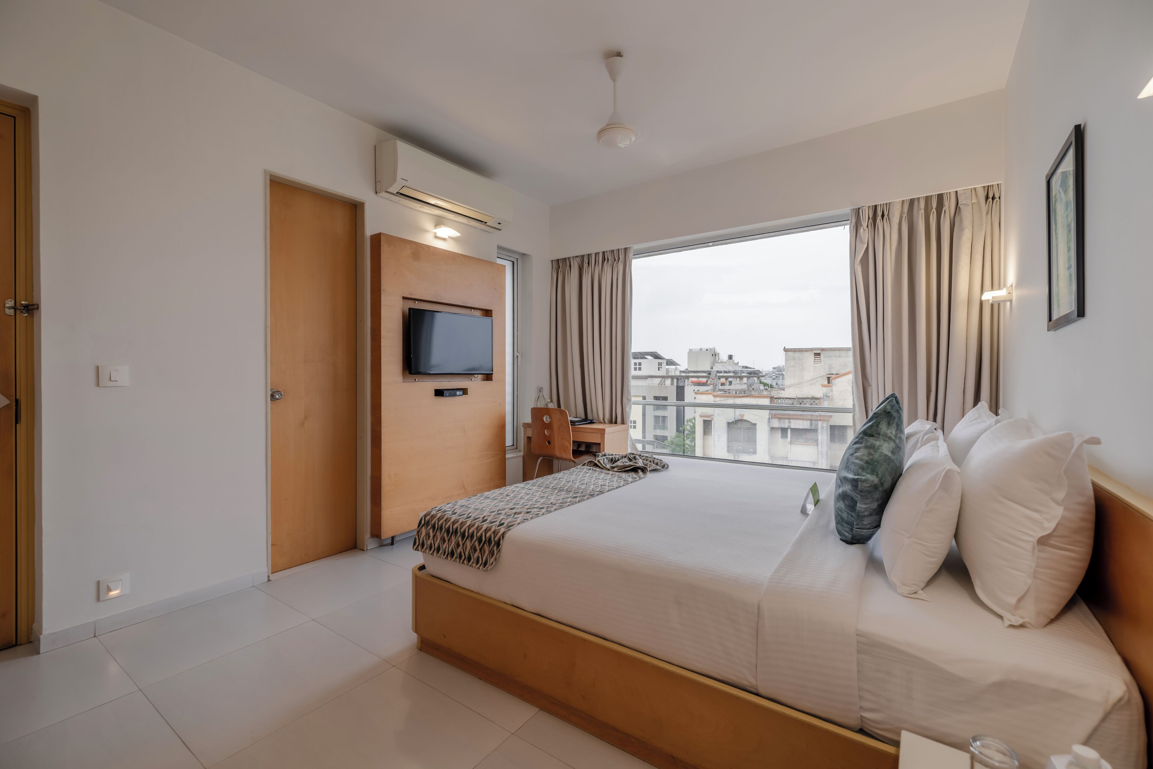 Habitación de hotel moderna con una cama grande, un televisor montado en la pared, un escritorio y un amplio ventanal con vistas a la ciudad.