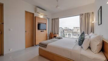Habitación de hotel moderna con una cama grande, un televisor montado en la pared, un escritorio y un amplio ventanal con vistas a la ciudad.