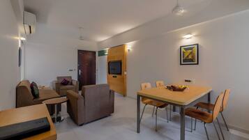 A modern living room with a sofa, a small table, and a dining area with a table and chairs. There is a TV mounted on the wall and a door leading to another room.