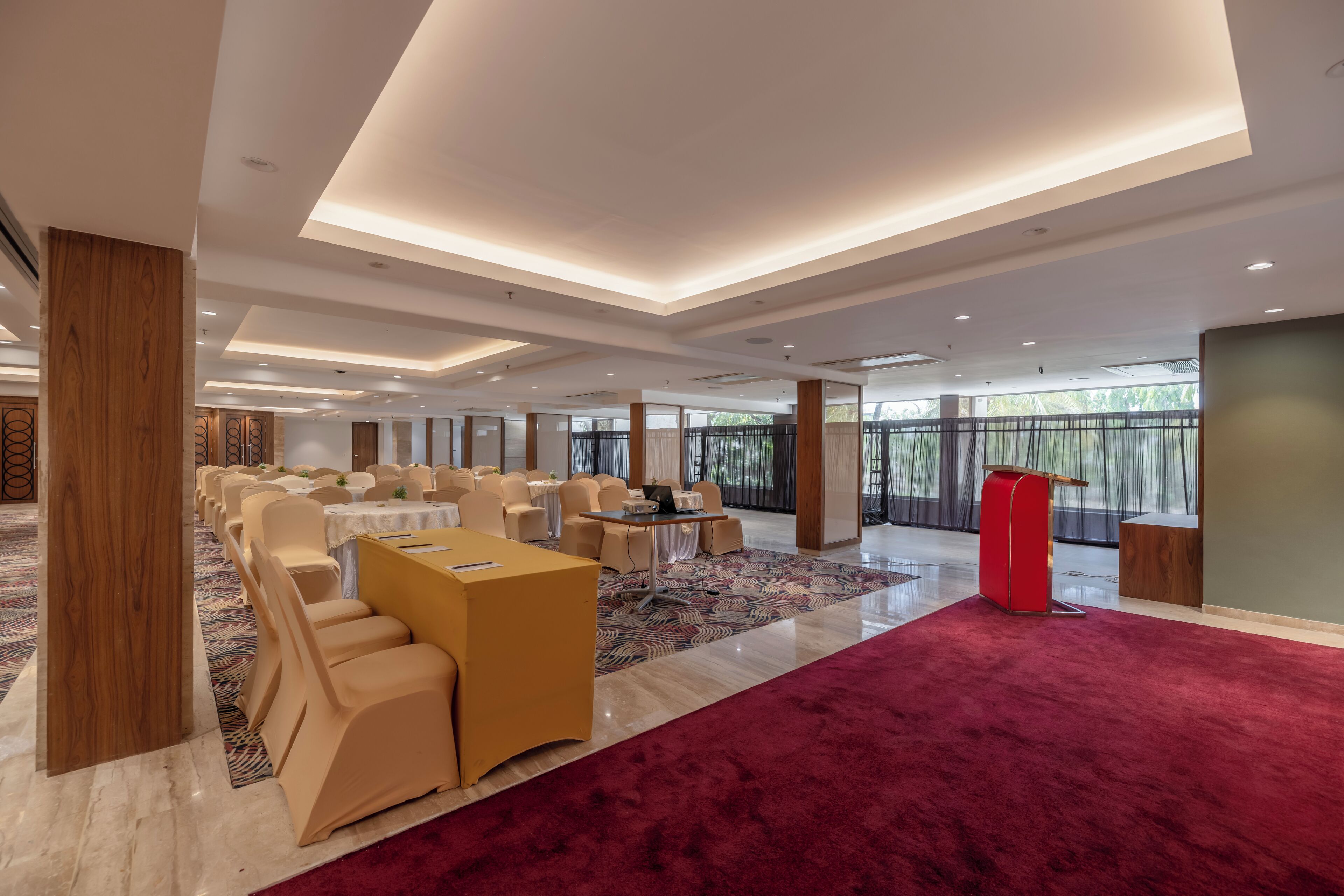 A spacious conference room with rows of chairs, a stage with a podium, and large windows offering a view of the outdoors.