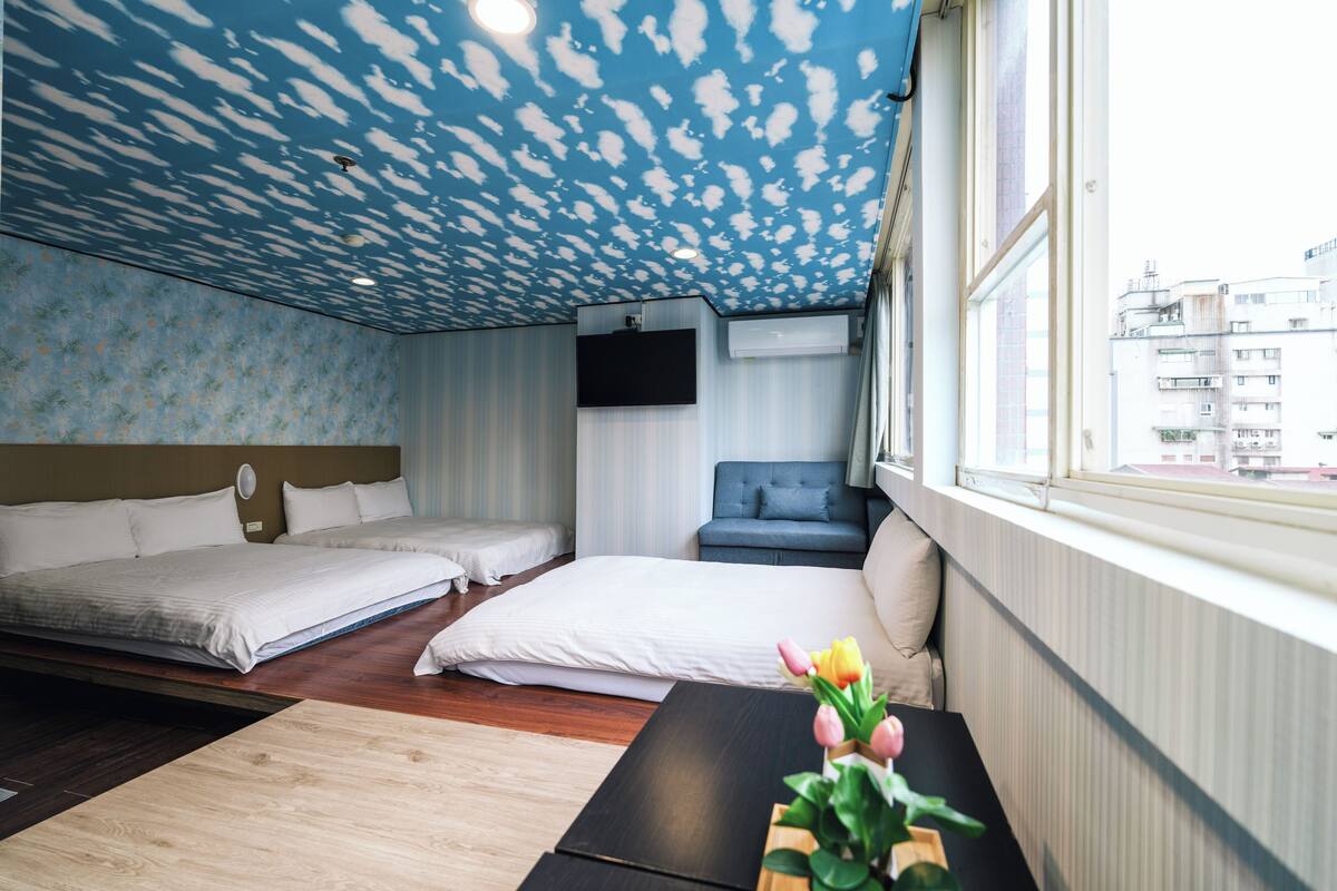 A hotel room with two beds, a TV, a window with a view of buildings, and a table with a flower vase.
