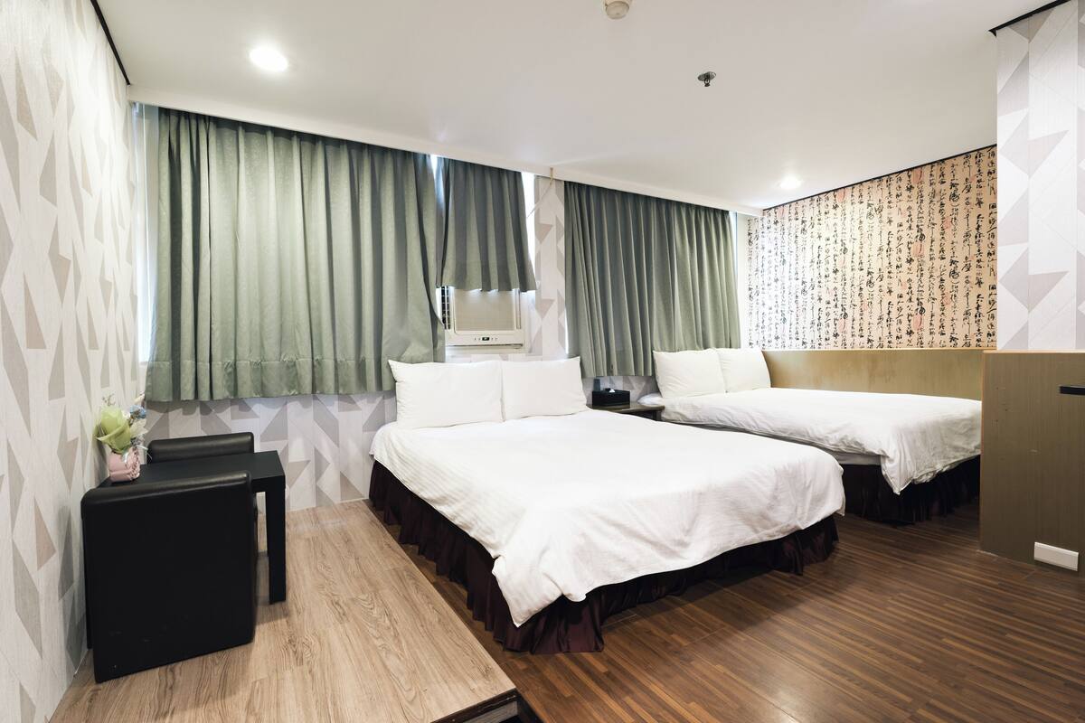 A hotel room with two beds, a small table, a bedside table, a wall with calligraphy, and a window with curtains.