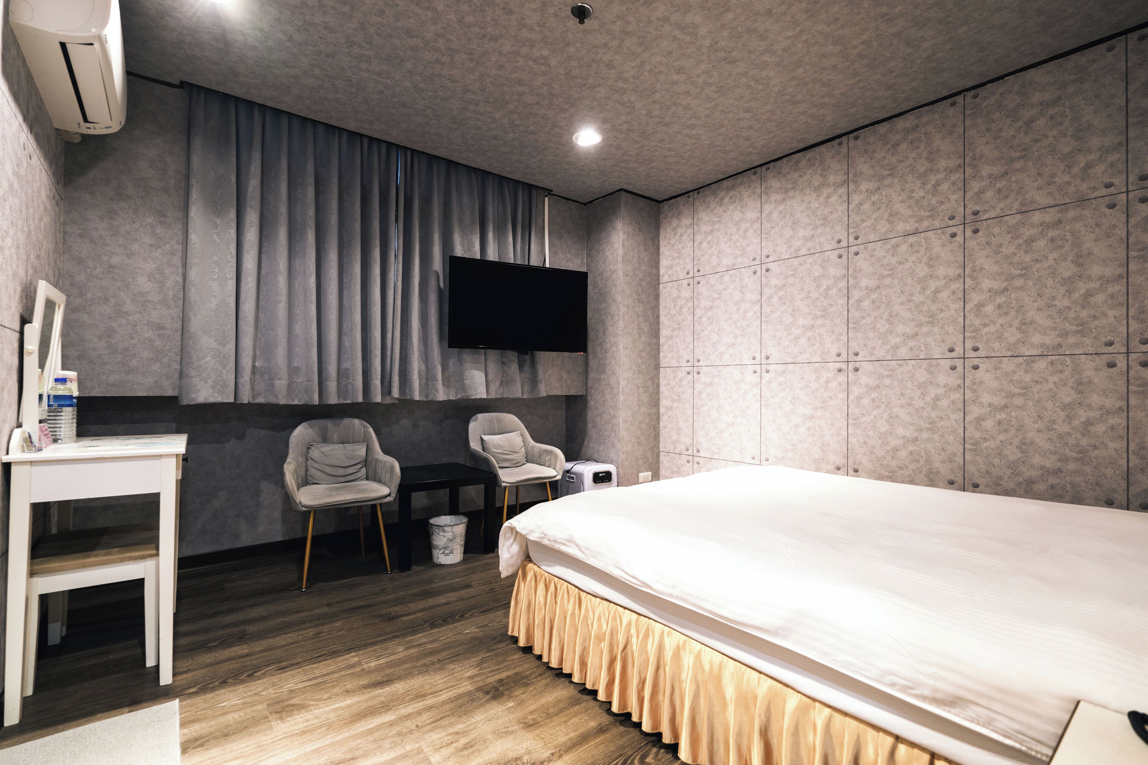 A modern hotel room with a large bed, two chairs, a desk, a flat-screen TV, and a wardrobe.