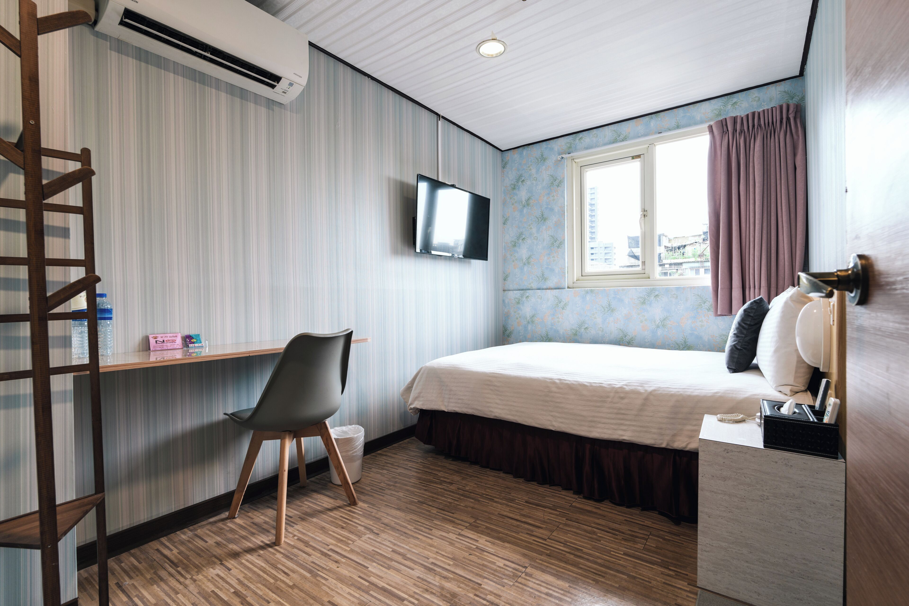 Ein Schlafzimmer mit Bett, einem an der Wand montierten Fernseher, einem Fenster mit Vorhängen, Holzboden und einem Schreibtisch mit Stuhl.