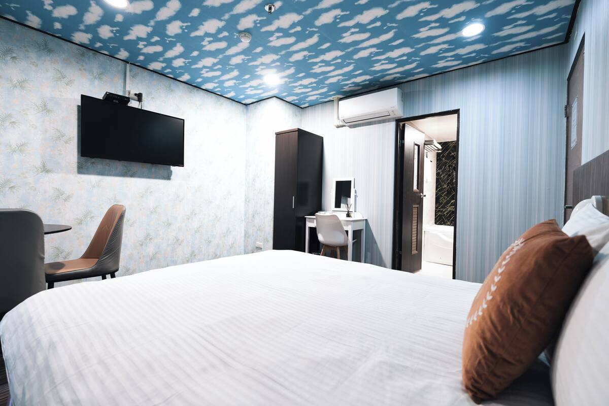 A modern hotel room with a large bed, a desk, a chair, and a TV mounted on the wall.