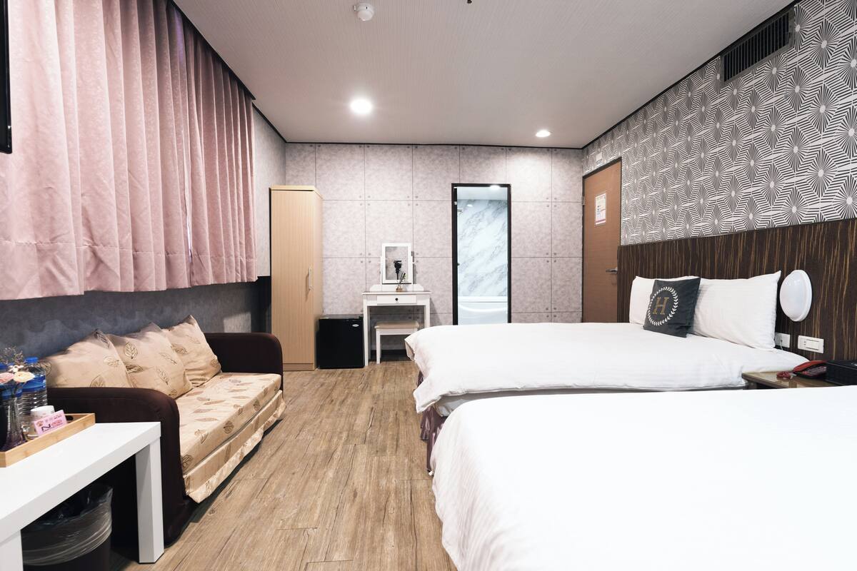 A modern hotel room with a large bed, a sofa, a desk, and a bathroom visible through a glass door.