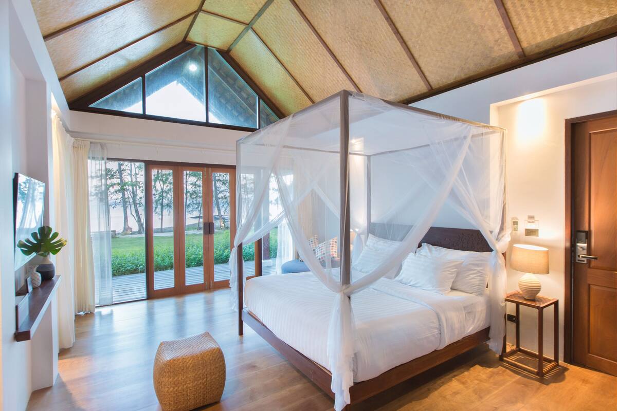 A bedroom with a four-poster bed, a bedside table with a lamp, and a view of the outdoors through large sliding doors.