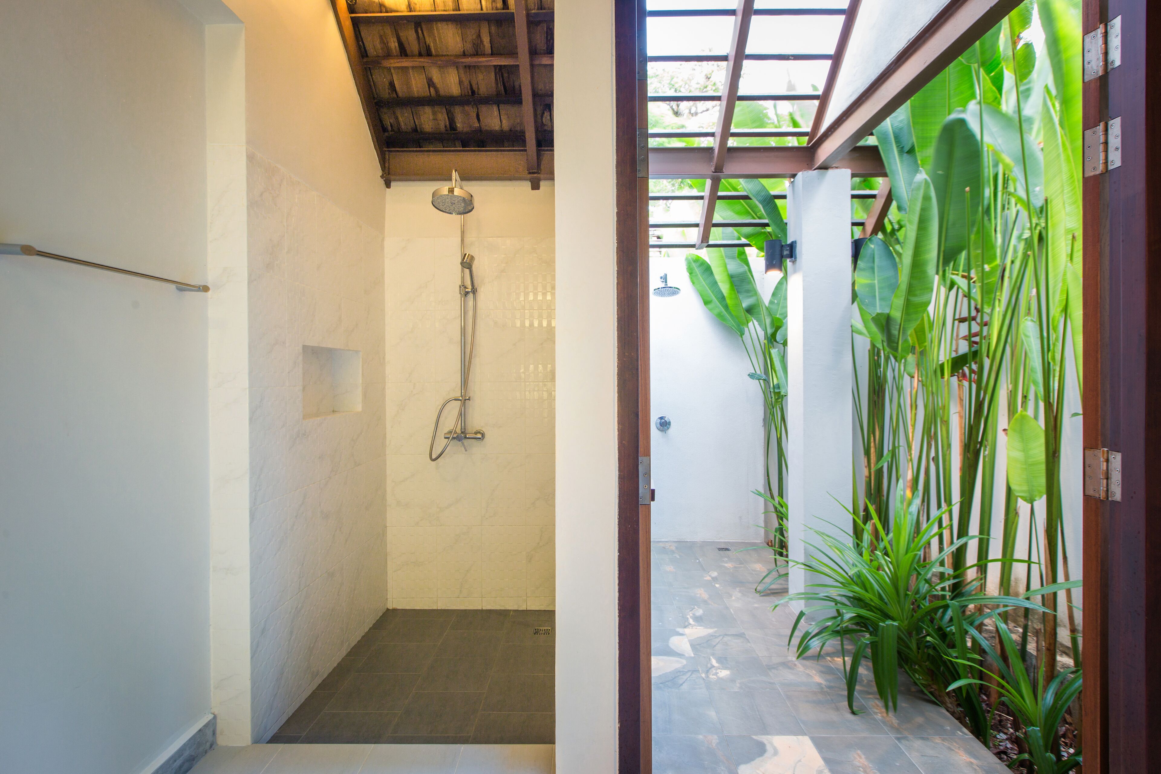 Casa de banho com chuveiro, chão em azulejo e vista para um jardim com folhas verdes grandes.