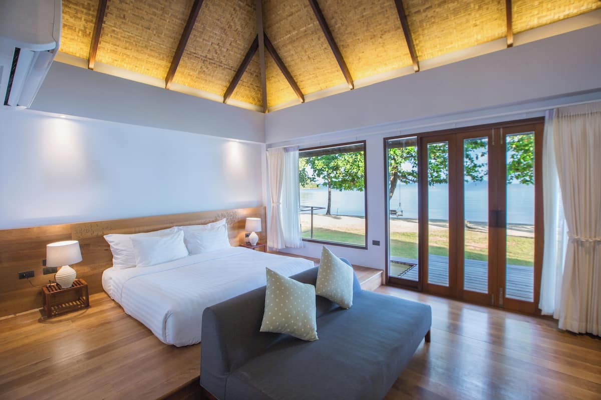 A bedroom with a bed, a sofa, a nightstand, and a window with a view of the beach.