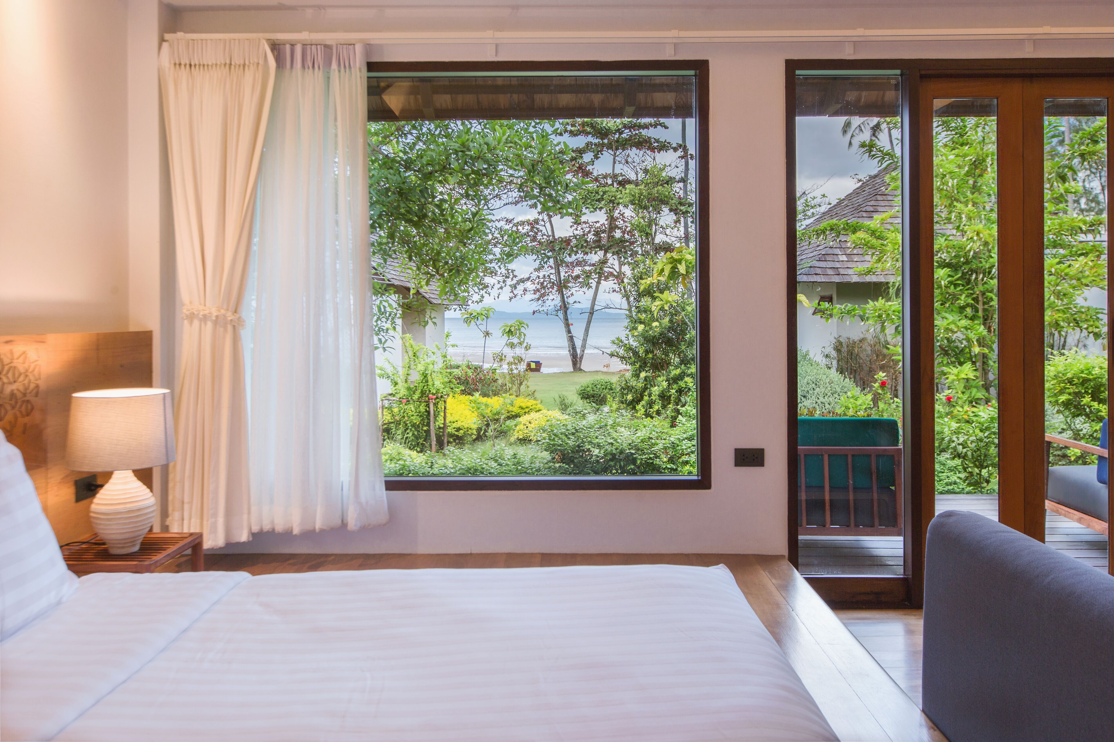 A bedroom with a large window offering a view of a garden and a building across the water.