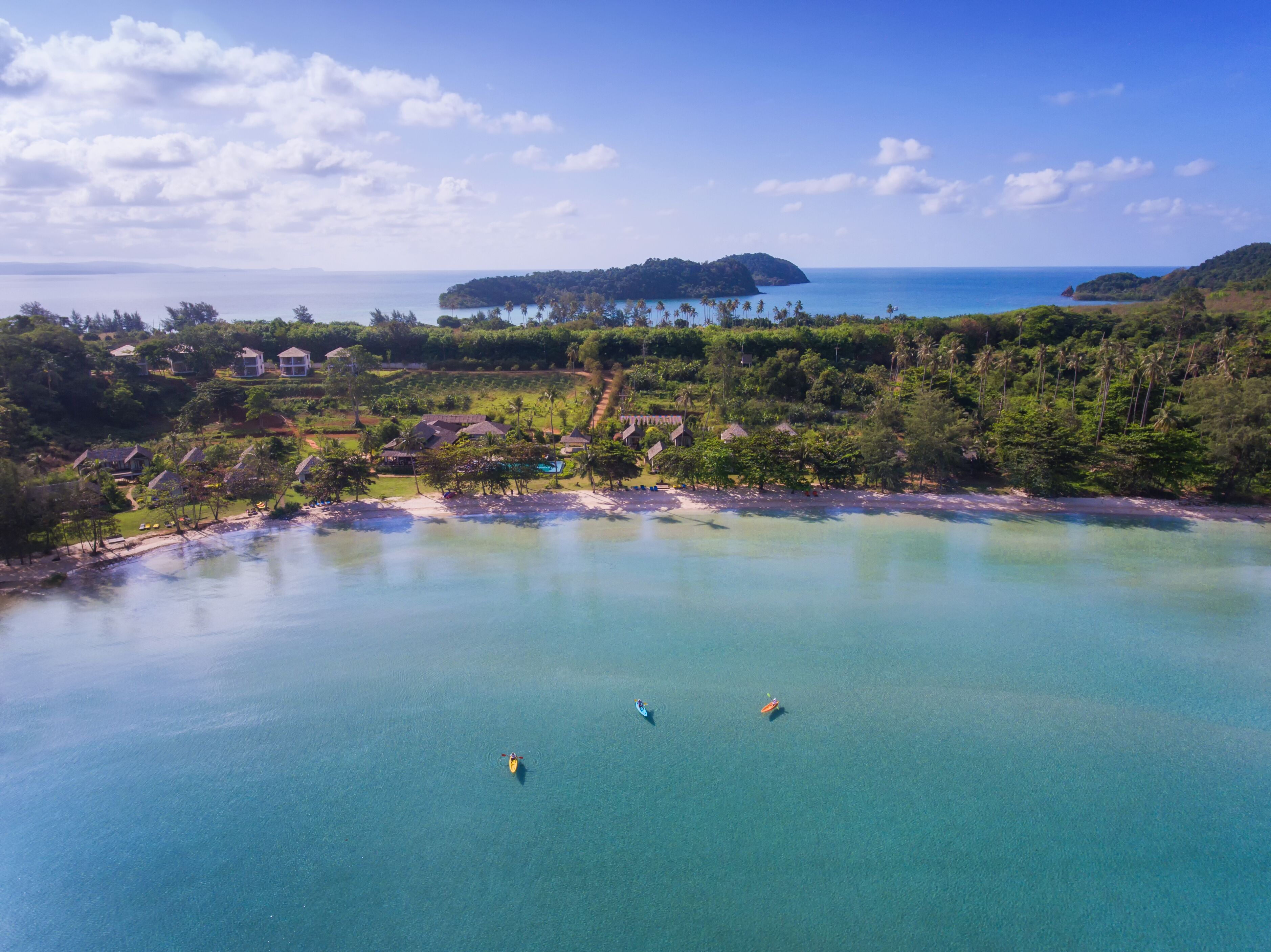 Une vue aérienne d’une zone côtière, avec une eau turquoise cristalline, une petite île au loin et une plage bordée de quelques maisons et de quelques kayaks.