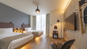 Habitación de hotel con dos camas, un escritorio, una silla, un televisor y un ventanal con cortinas.