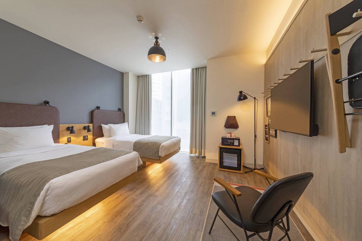 Une chambre d’hôtel avec deux lits, un bureau, une chaise, une télévision et une grande fenêtre avec des rideaux.
