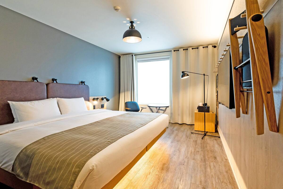 Une chambre d’hôtel avec un grand lit, un bureau, une chaise, une télévision et une fenêtre avec des rideaux.