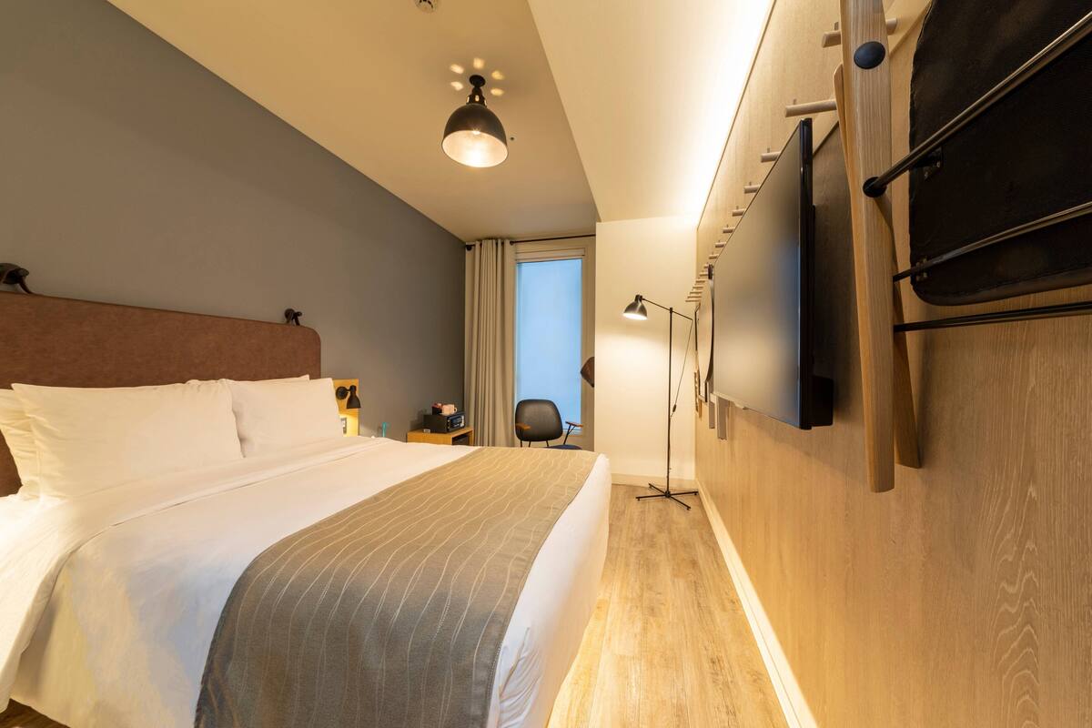 Une chambre d’hôtel avec un grand lit, un bureau, une chaise, un téléviseur fixé au mur et une fenêtre avec des rideaux.