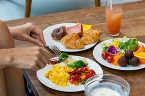 Une table en bois sur laquelle sont disposés des plats contenant des œufs, du jambon, des croissants, des légumes et un verre de jus de fruits.