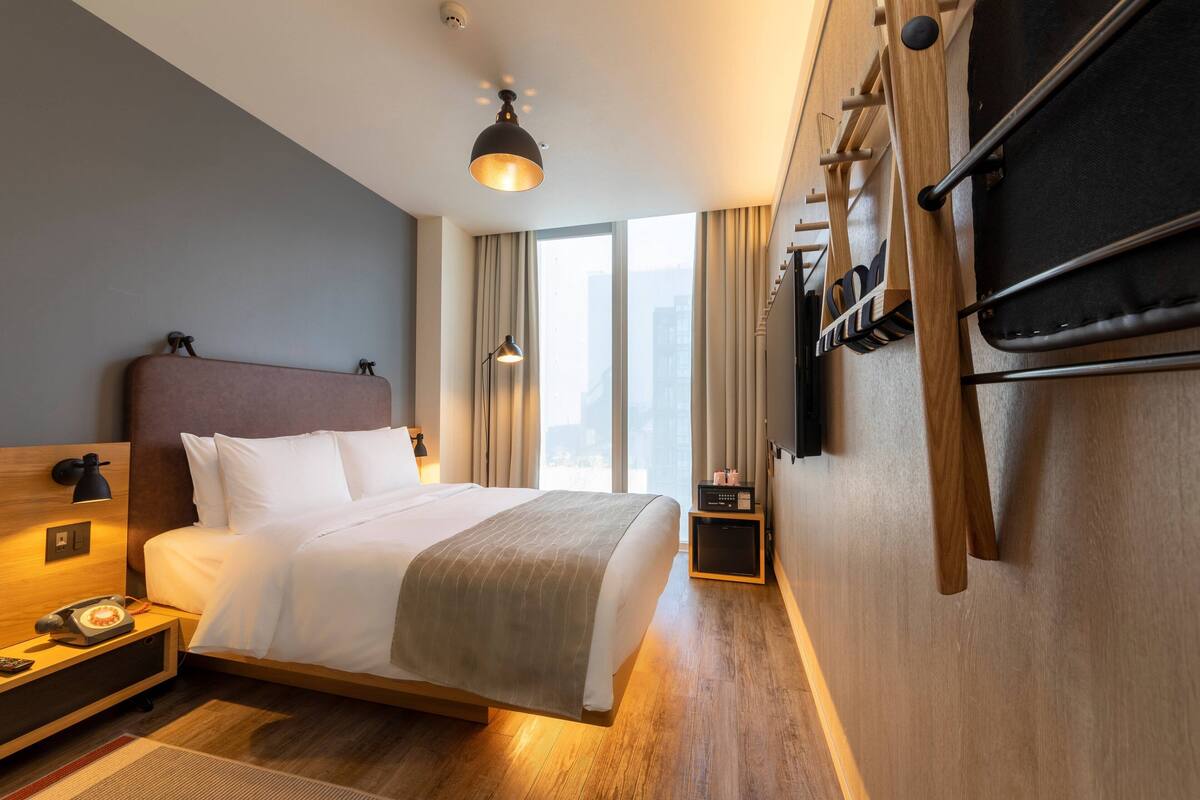 Une chambre d’hôtel moderne dotée d’un grand lit, d’un bureau avec un téléphone, d’une lampe et offrant une vue sur la ville grâce à de grandes fenêtres.