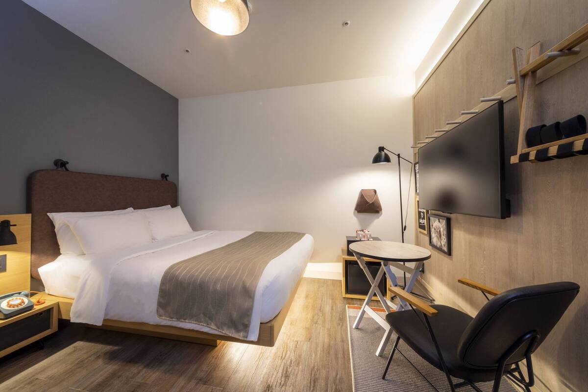 Une chambre d’hôtel moderne, équipée d’un lit, d’une table de chevet, d’une lampe, d’une chaise et d’un téléviseur fixé au mur.