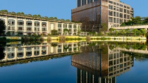 Un immeuble moderne de grande hauteur, doté d’une façade en verre, jouxtant un bâtiment historique aux fenêtres en arc de cercle, dont le reflet se mire dans une étendue d’eau calme.