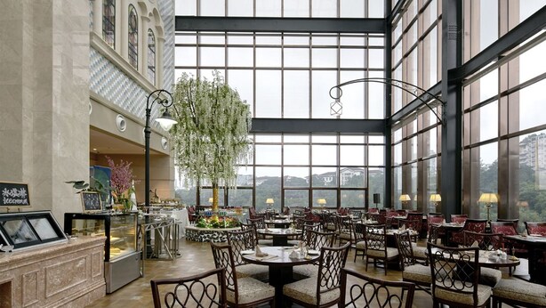 Un vaste espace intérieur aménagé en salle à manger, doté de hauts plafonds et de grandes fenêtres, avec un arbre central et de nombreuses tables et chaises.