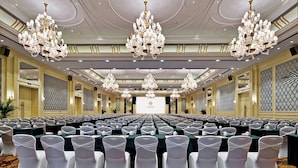 Une grande salle de conférence avec de longues rangées de chaises et de tables blanches, des lustres décoratifs et une moquette à motifs.
