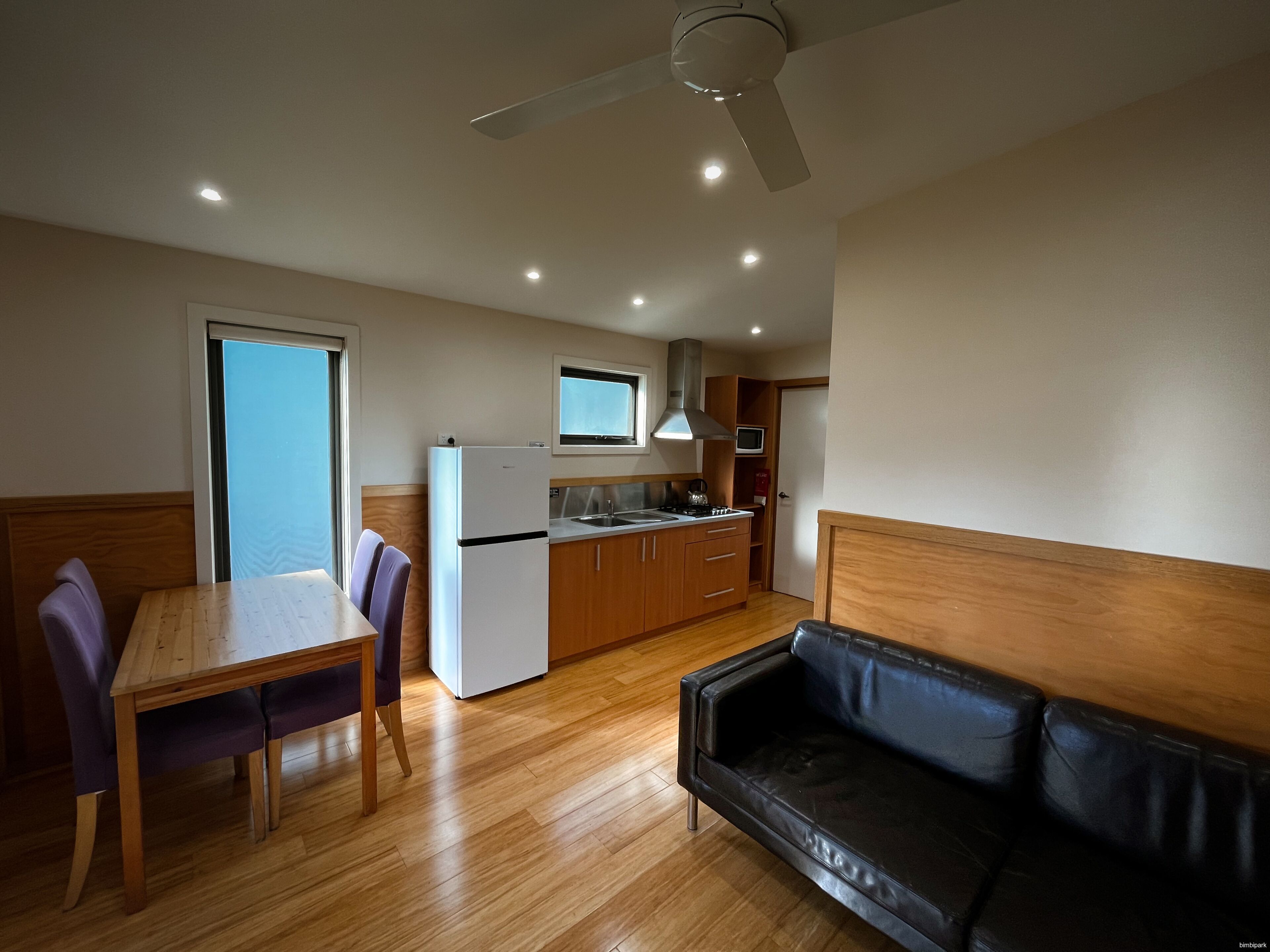 A compact kitchen with a refrigerator, stove, and microwave, a dining table with chairs, and a black leather sofa.