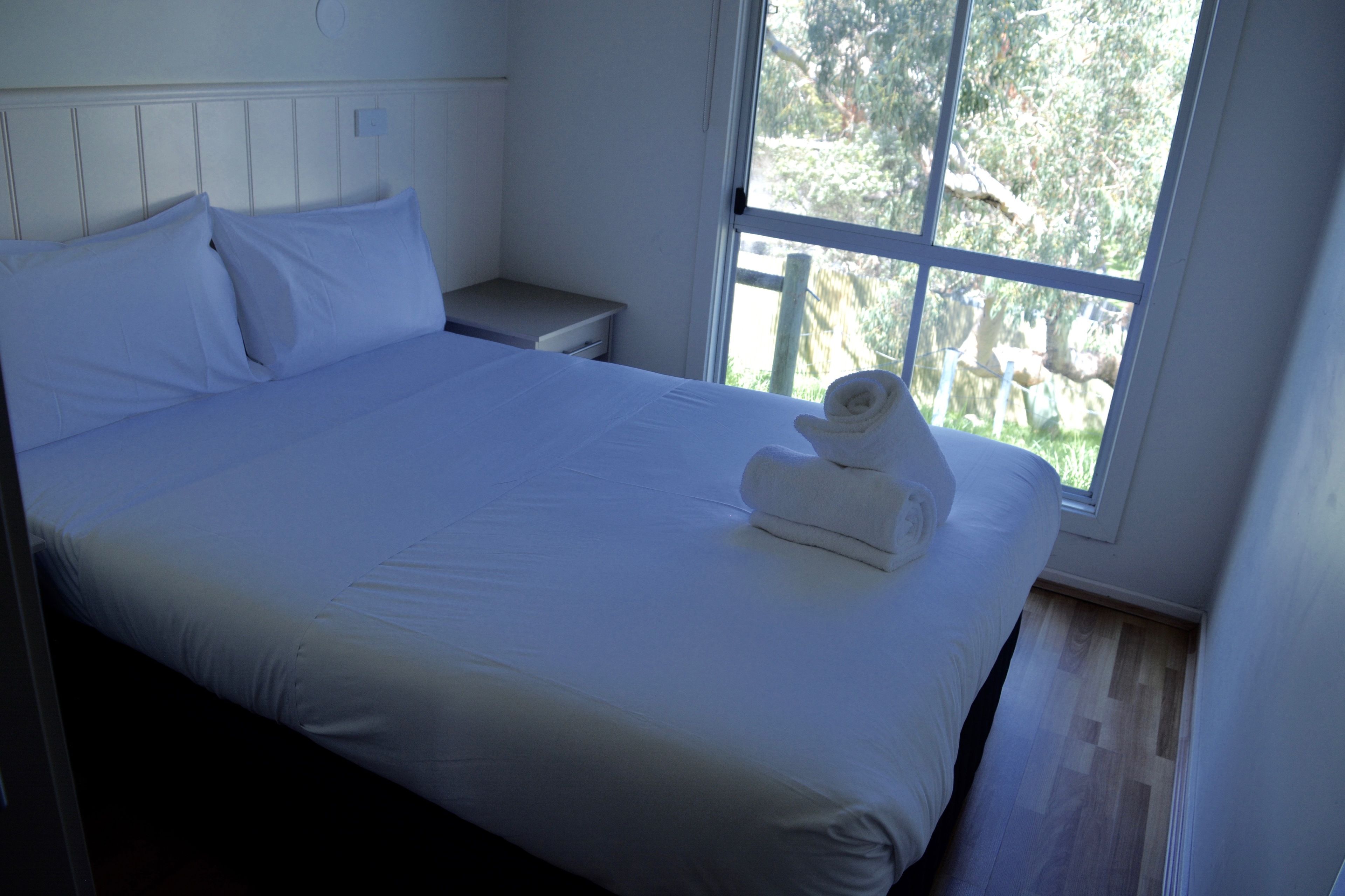 A neatly made bed with white linens and a folded towel, a bedside table, and a window with a view of greenery.