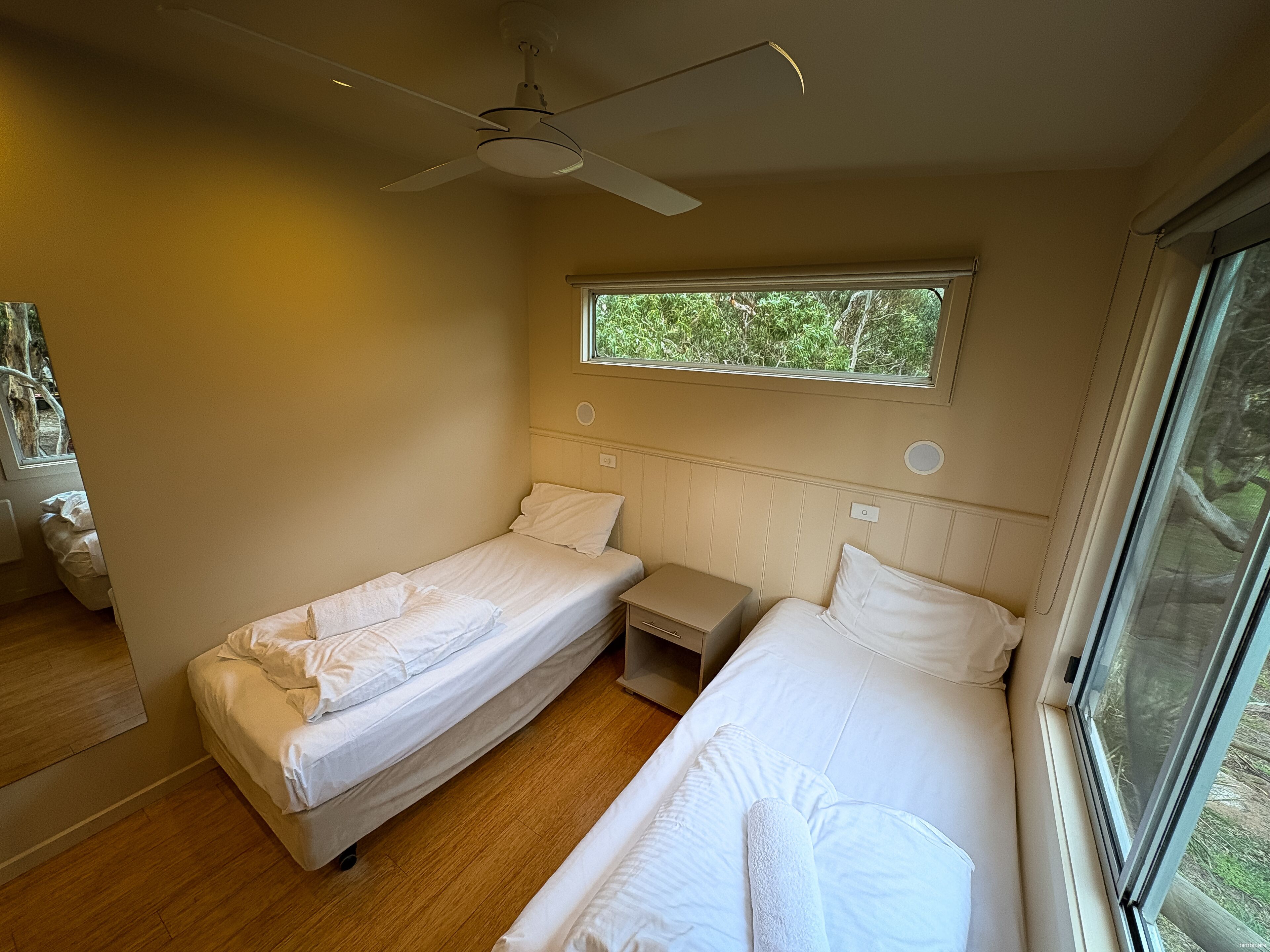 A room with two single beds, a ceiling fan, a window with a view of greenery, and a wooden floor.