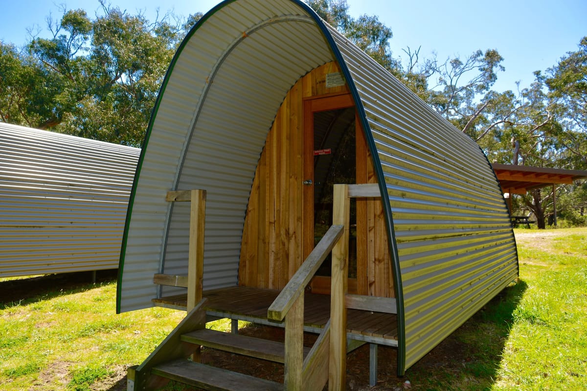 A metal and wooden RV with a ramp leading to the entrance, surrounded by grass and trees.