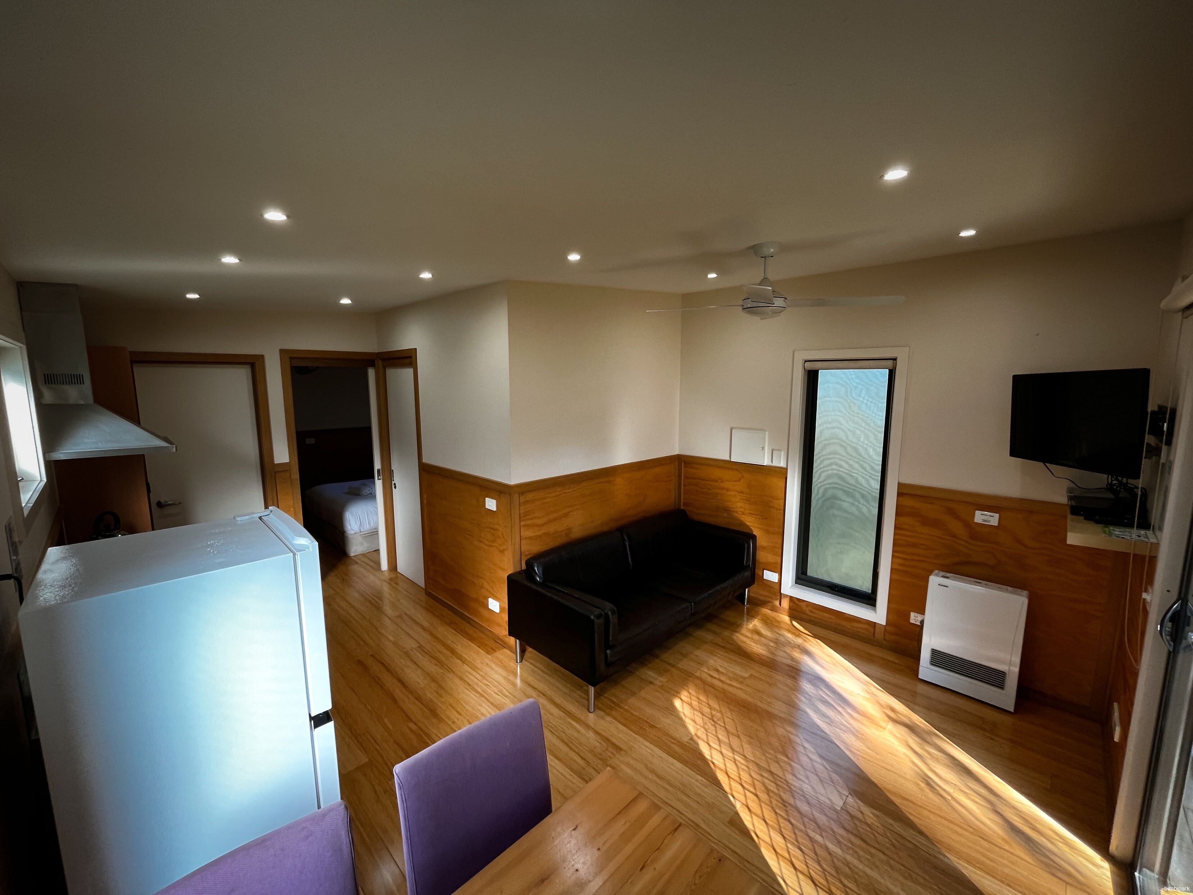 Un espace de vie compact comprenant un coin cuisine, un canapé noir, un fauteuil violet, une télévision et une grande fenêtre.