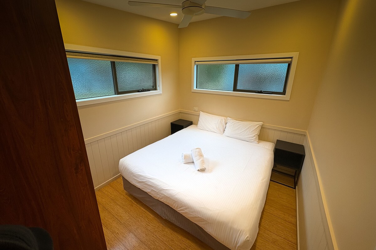 Une chambre à coucher comprenant un lit, deux tables de chevet, un ventilateur de plafond et des fenêtres à vitres dépolies.