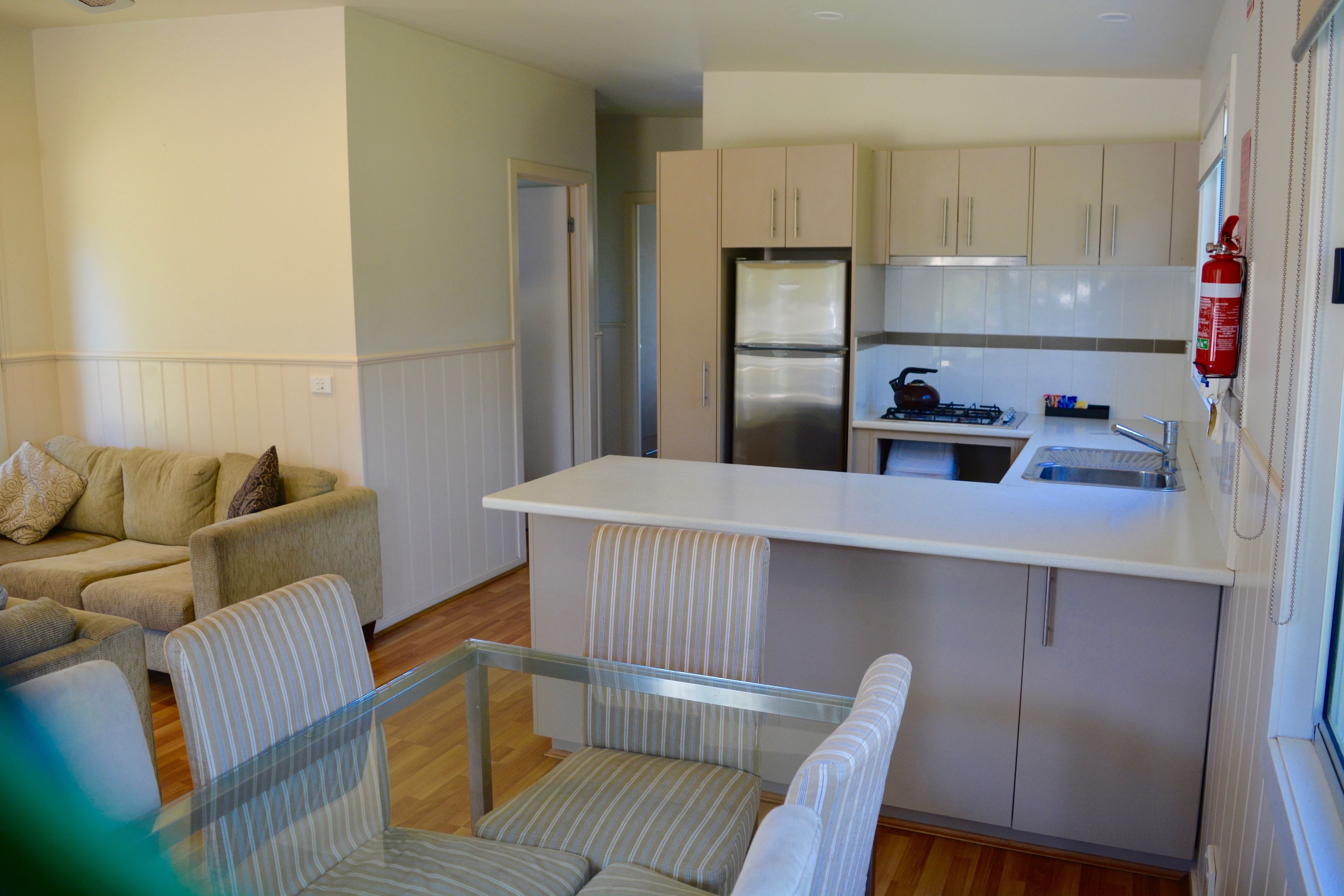 A compact kitchen with a refrigerator, stove, and sink. There is a dining area with a glass table and striped chairs. A sofa is visible in the background.