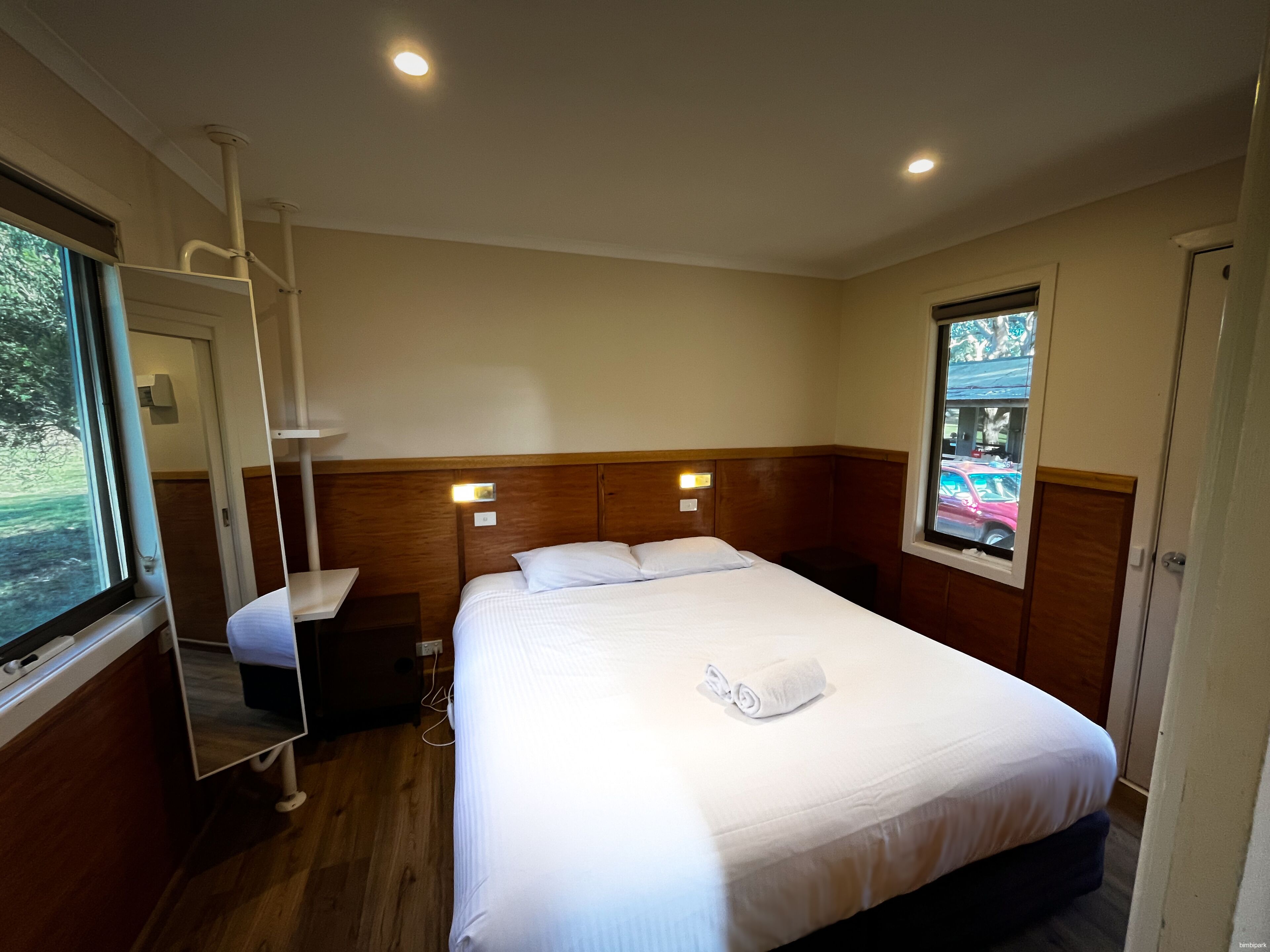 A hotel room with a single bed, a bedside table, a mirror, a window with a view of the outside, and a wooden headboard.
