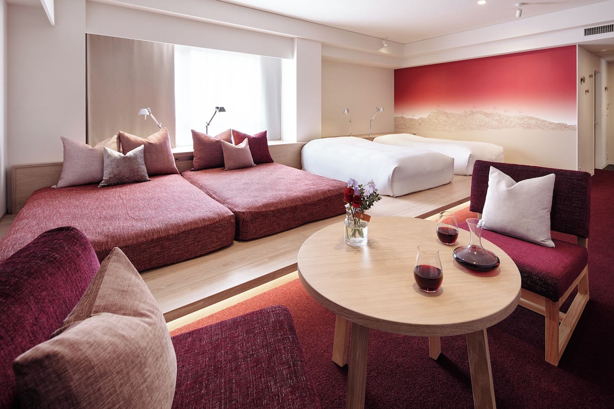 Una habitación de hotel moderna con una cama grande, un sofá, una mesa de comedor con copas de vino y una pared decorada en rojo.